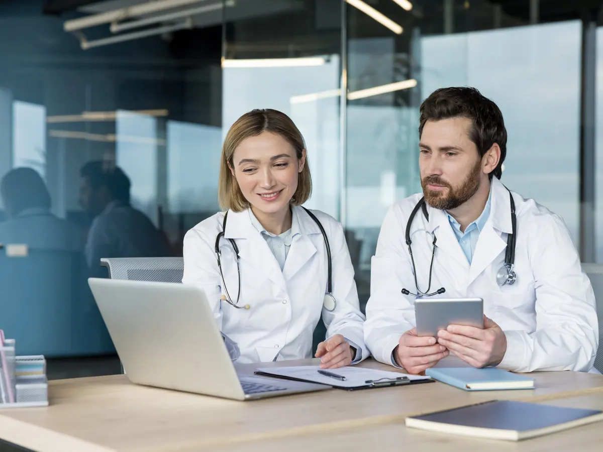 Two clinicians in white coats reviewing information on a laptop and a tablet at a desk in a bright office