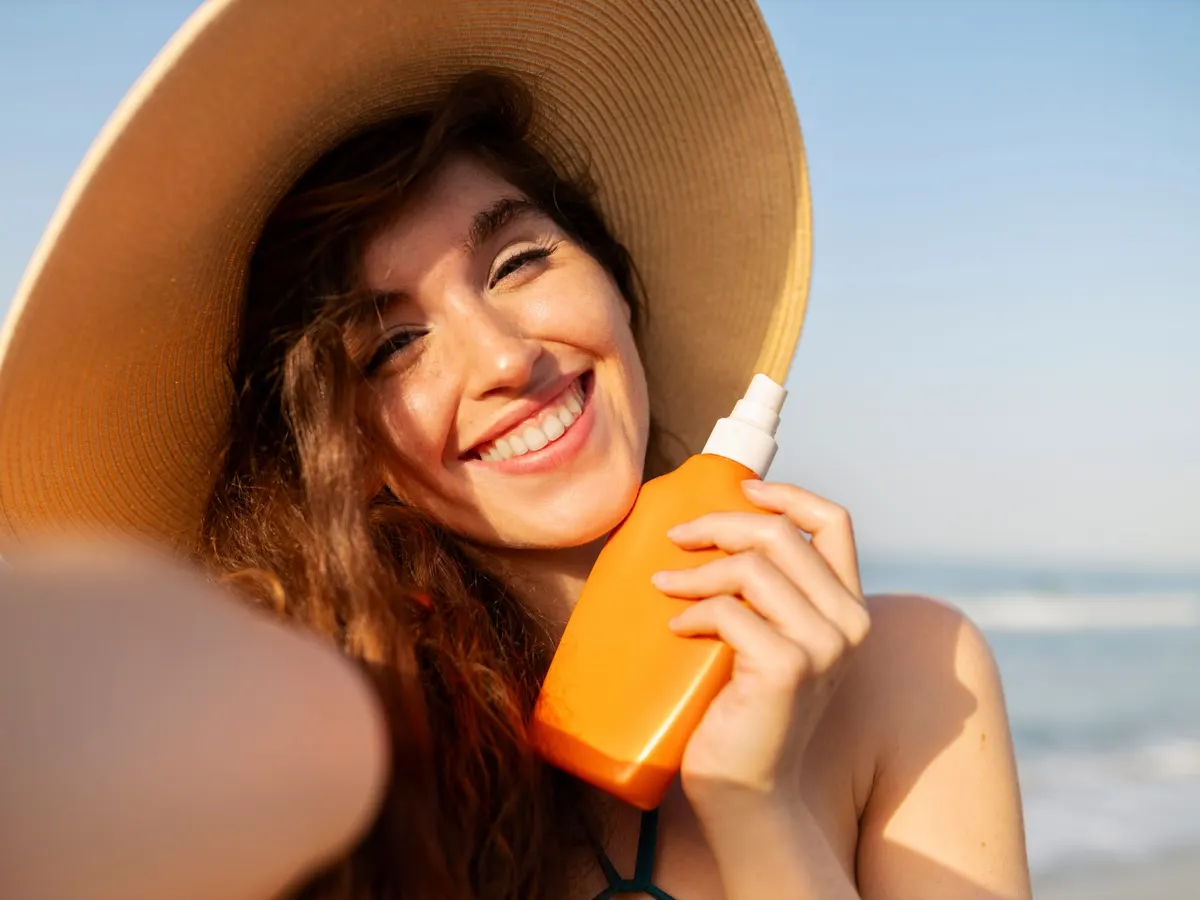 Smiling woman on a sunny beach holding a bottle of sunscreen