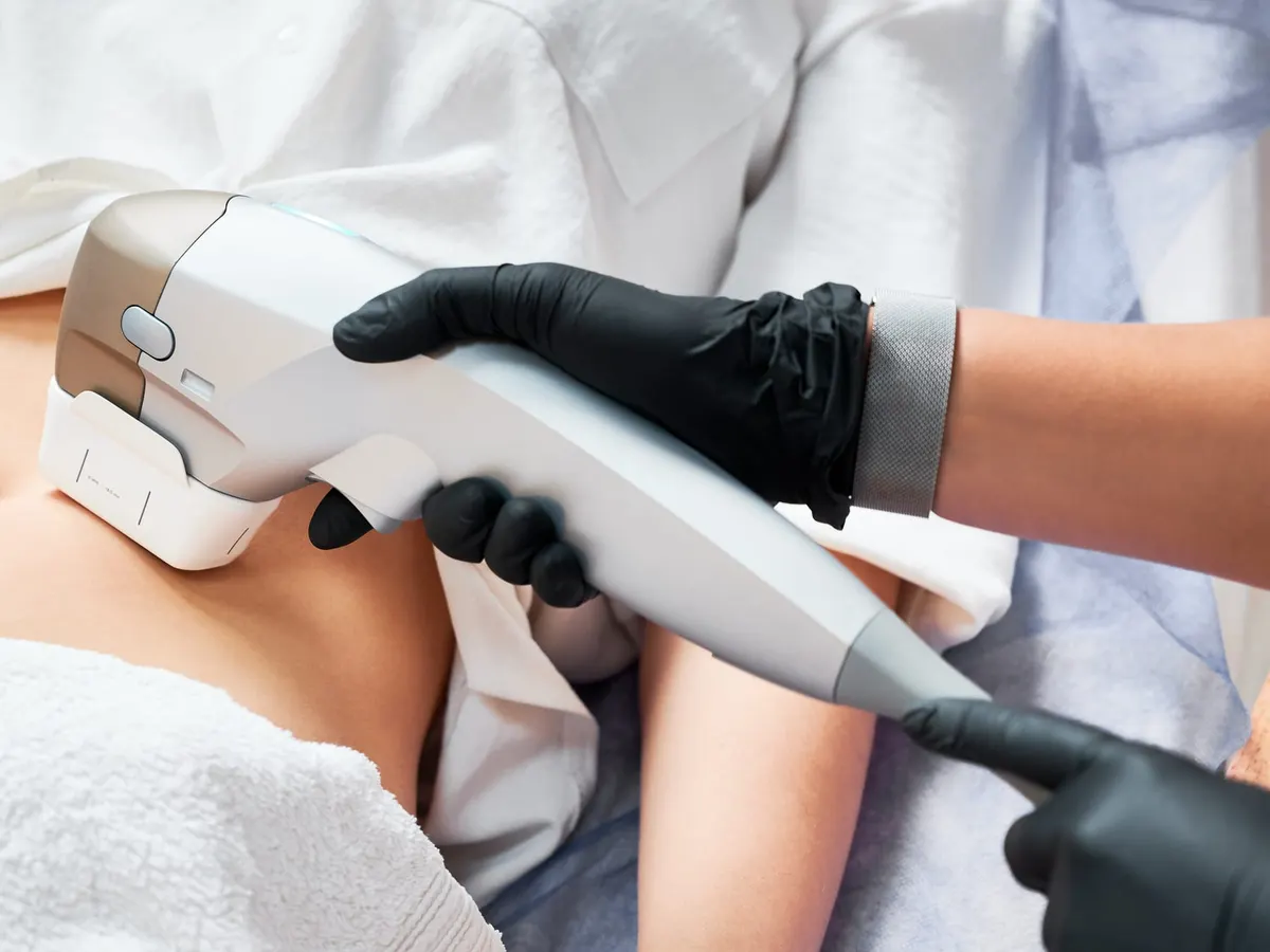 Gloved practitioner holding an ultrasound handpiece over a draped patient's abdomen during a body contouring session