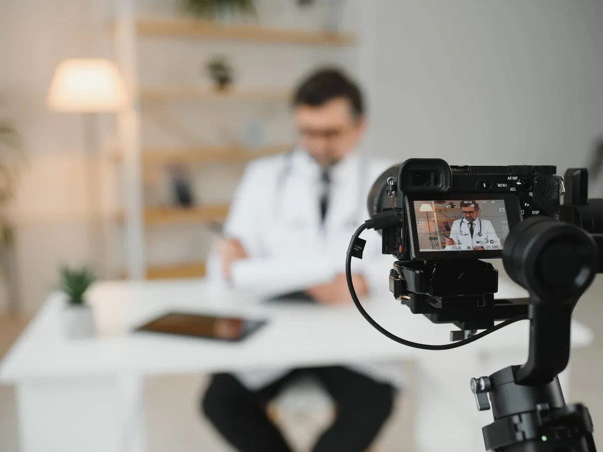 A doctor in a white coat seated at a desk being filmed by a camera on a handheld stabiliser in a bright office