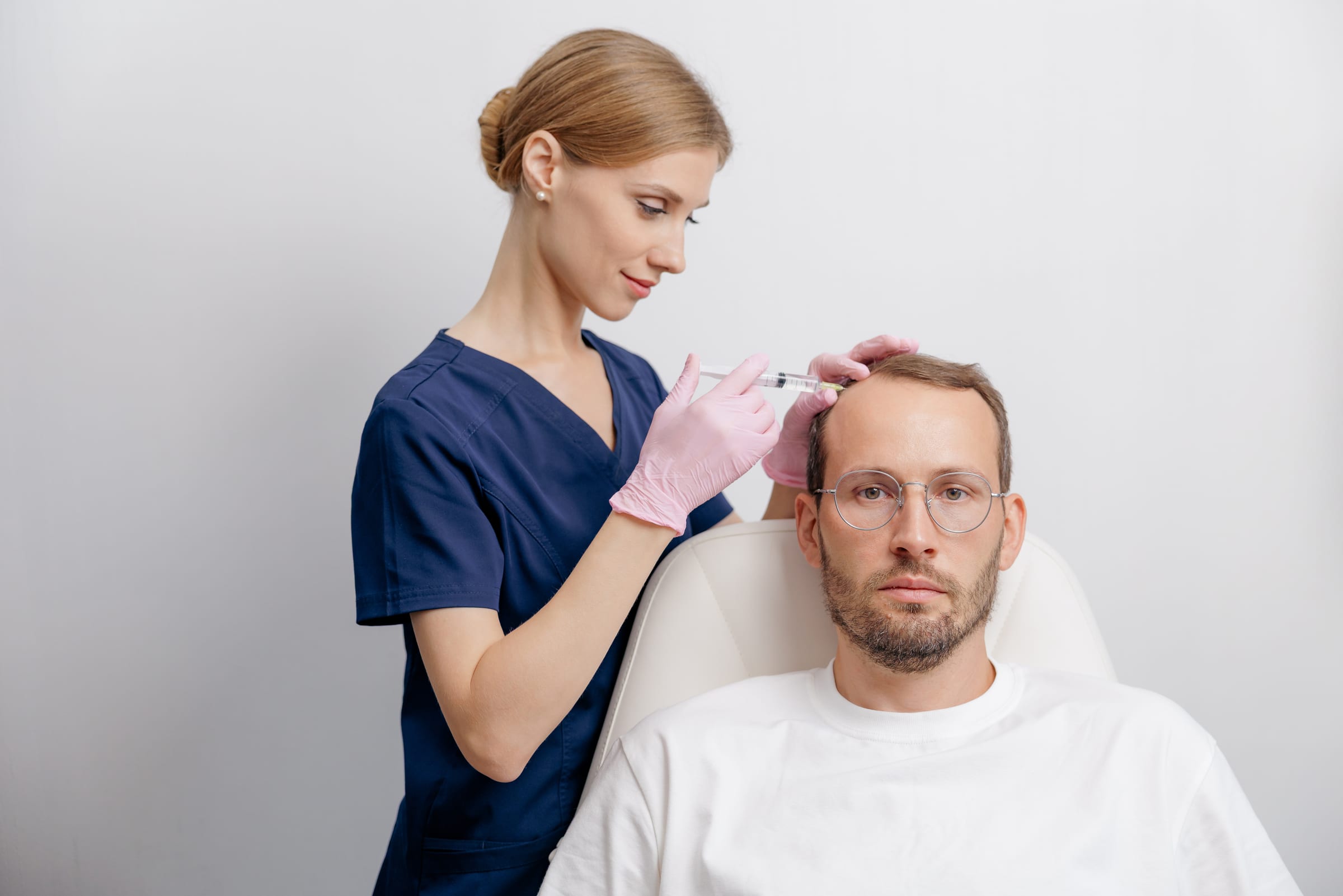 Trichologist in blue scrubs administering a scalp injection to a seated male patient in a bright clinic